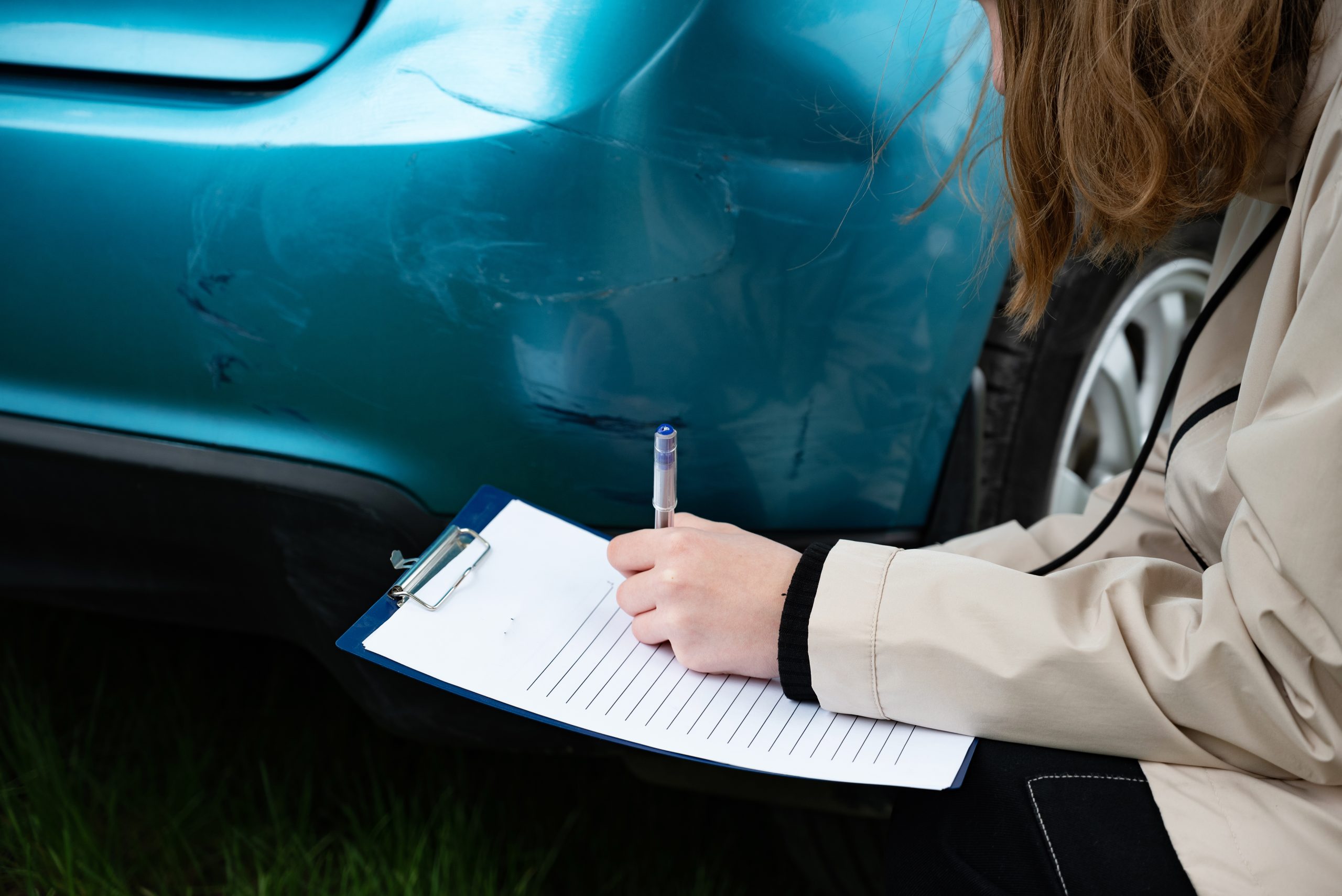 Person inspecting car bumper damage with clipboard outdoors.