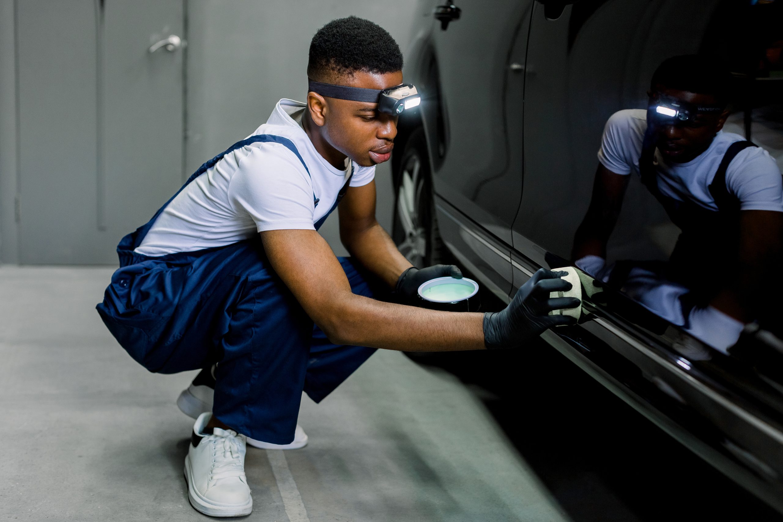 Car detailing and polishing concept. Young African man worker, wearing uniform and gloves, holds the sponge in hand and polishes the car body with solid wax. Selective focus.