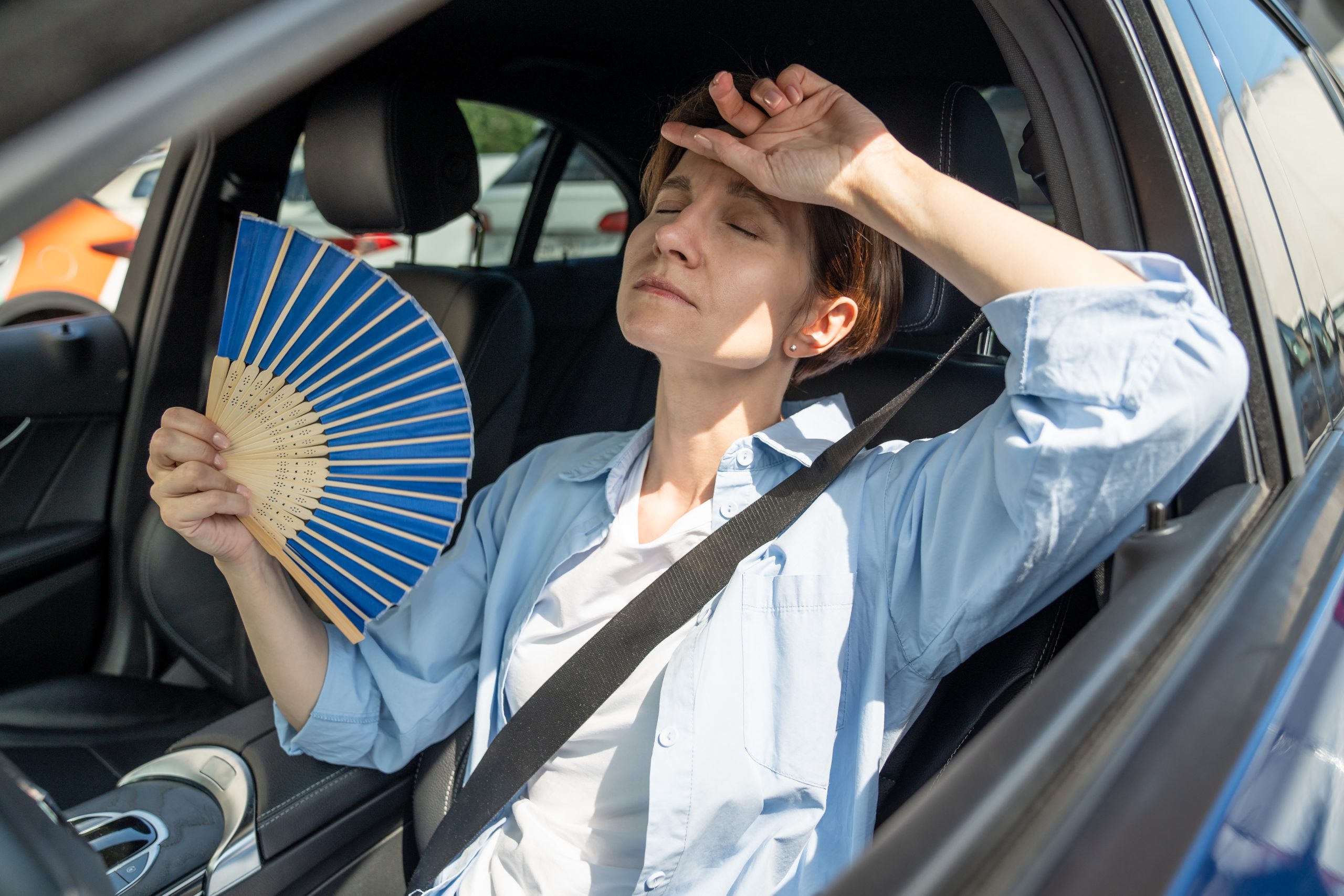 Exhausted middle aged woman drives car waves blue fan suffers from stuffiness in traffic jam.