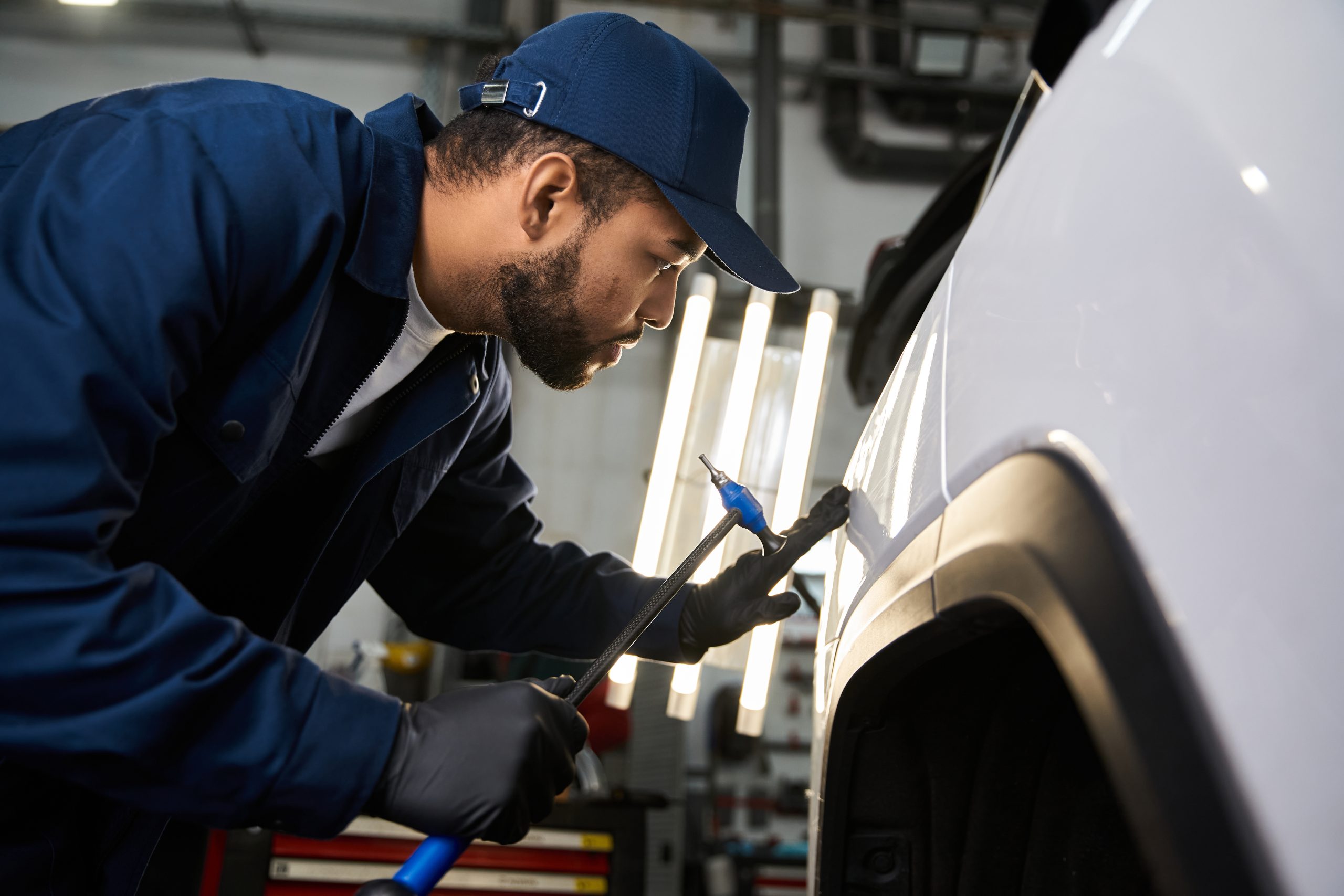 A focused young mechanic repairs a car in a bustling, fluorescent lit workshop.